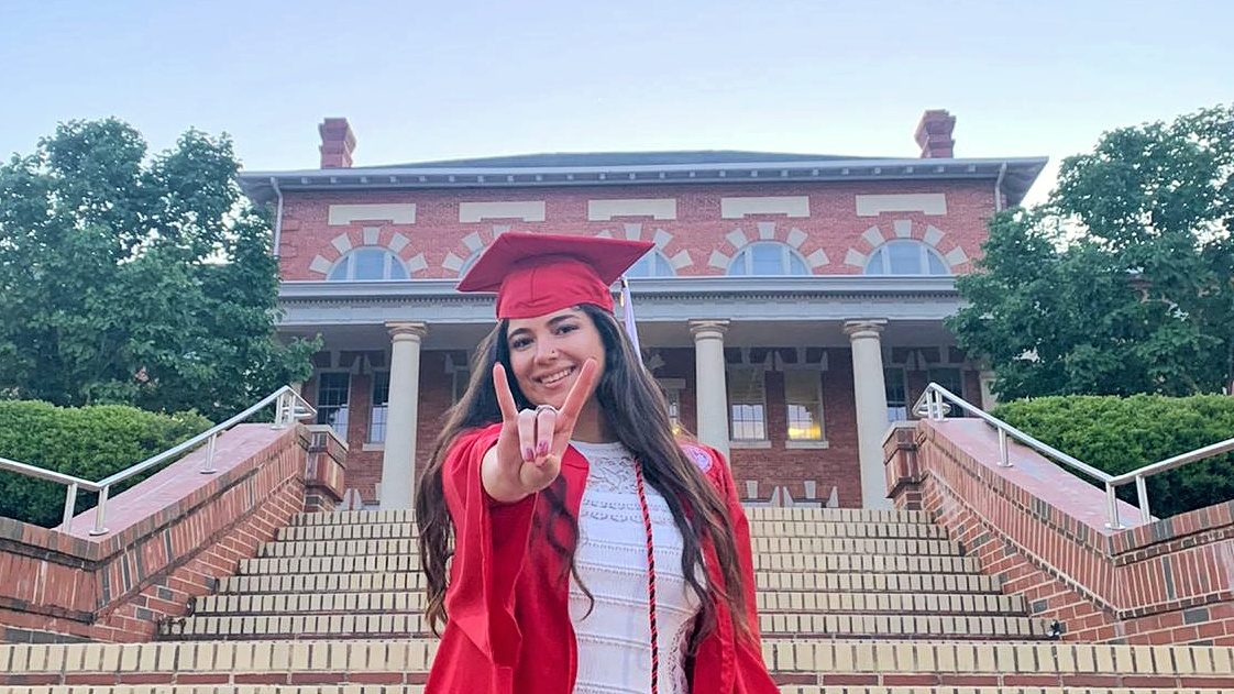 Esmira Poladova does the wolfie in front of a campus building in her cap and gown