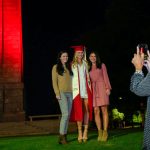 A woman takes a photo of three other women, one a new graduate, in front of the Belltower lit red.